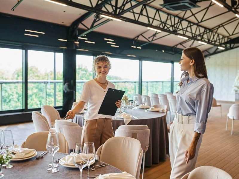 event-organizer-with-clipboard-pointing-at-table-with-festive-setting-near-woman-in-banquet-hall.jpg